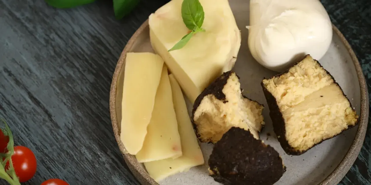 Różnorodne rodzaje serów na talerzu: kawałki żółtego sera, kulka mozzarelli i ser w ciemnej skórce.