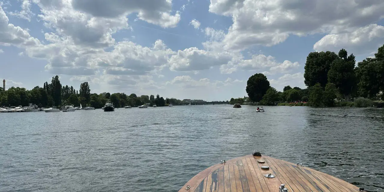 Blick vom Bug eines Holzbootes auf einen Fluss mit vielen Booten und grünen Ufern. Ein schöner Tag, fast wie ein Heidelberg See.