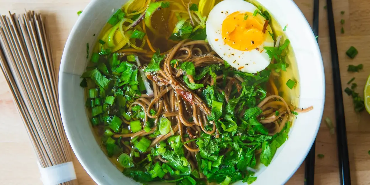 A miseczce aromatyczny rosół japoński z makaronem soba, jajkiem na pół i zieloną cebulką. Obok leżą pałeczki i surowy makaron.