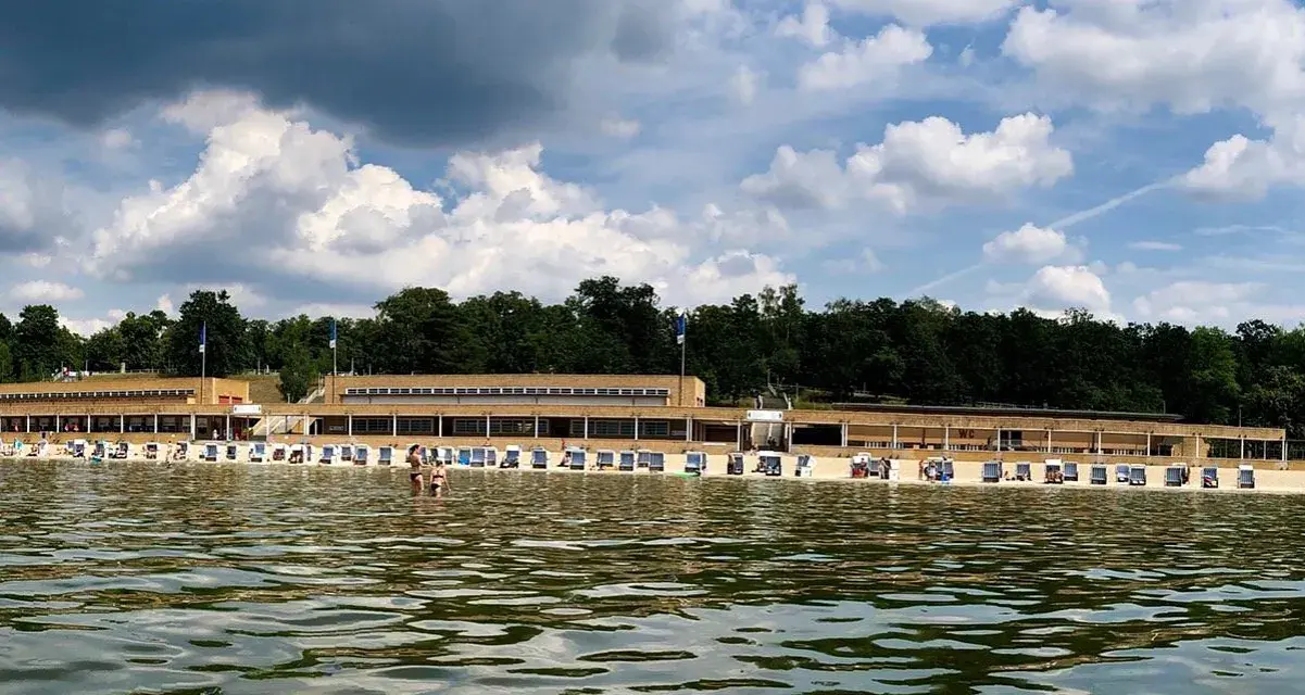 Das Strandbad Wannsee mit seiner langen Promenade und vielen Liegestühlen, im Hintergrund grüne Bäume und ein bewölkter Himmel.