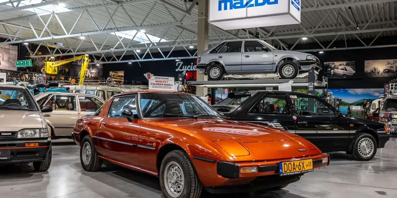 Pomarańczowa Mazda RX-7 błyszczy w muzeum samochodów, obok innych klasyków marki.