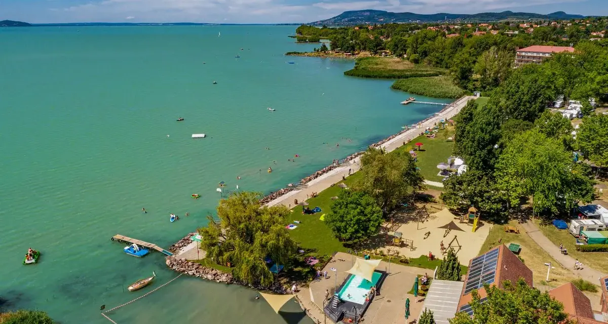 Słoneczny dzień na kempingu Balaton. Ludzie kąpią się w jeziorze i basenie, bawią na placu zabaw, a kampery stoją wśród drzew.