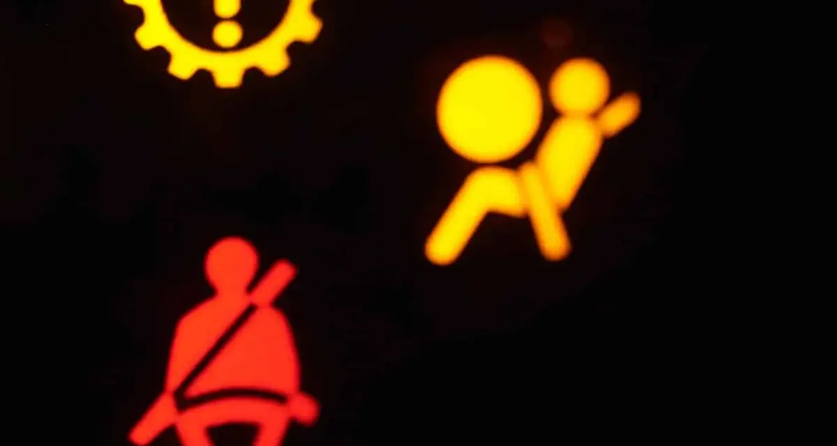 Das gelbe airbag symbol leuchtet. Daneben ein rotes Symbol für den Sicherheitsgurt. Ein gelbes Zahnrad mit Ausrufezeichen ist oben links zu sehen.