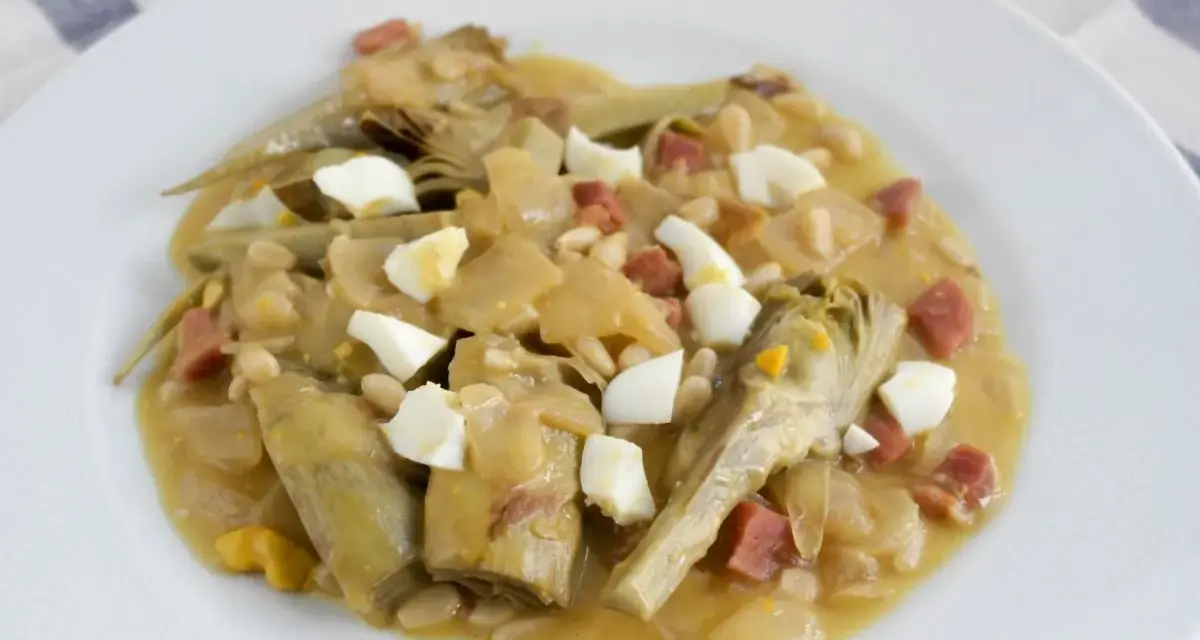 Plato de alcachofas con jamón, huevo duro y piñones, servido en una cazuela.