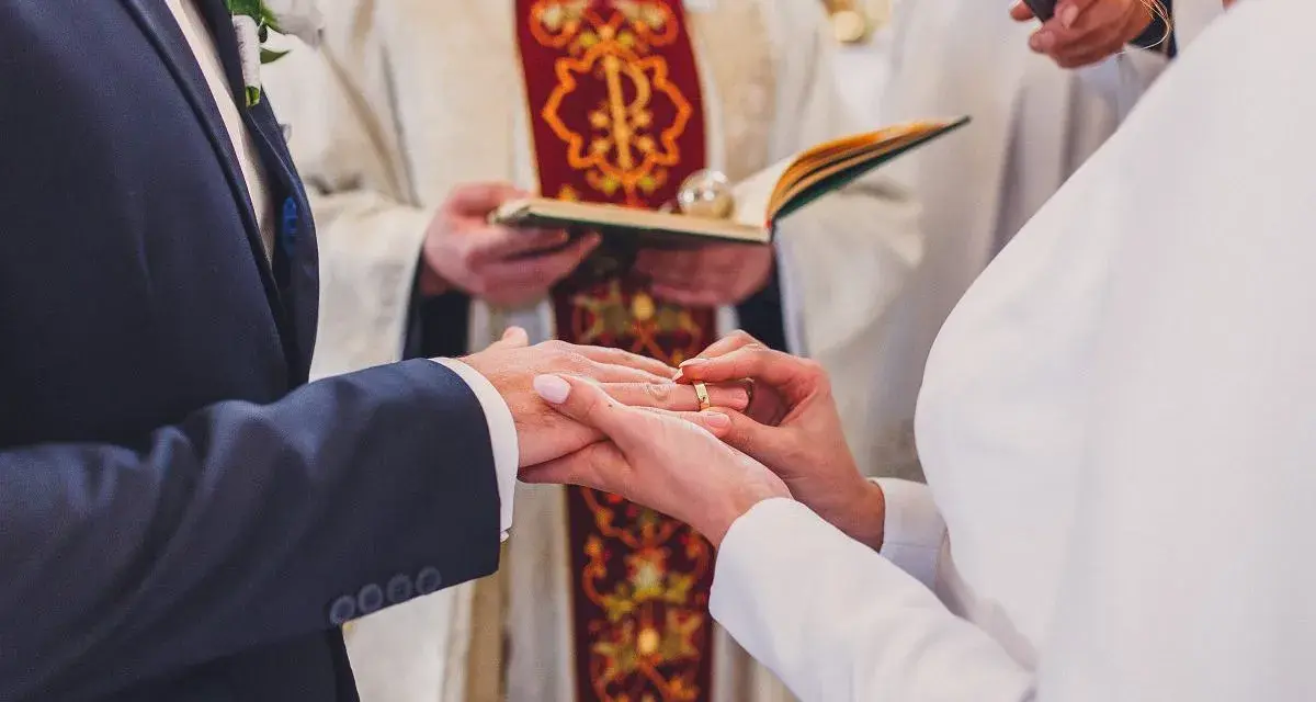 Ceremonia ślubna w kościele. Młoda para wymienia obrączki. W tle ksiądz z księgą. Warto wiedzieć, ile kosztuje ślub kościelny.