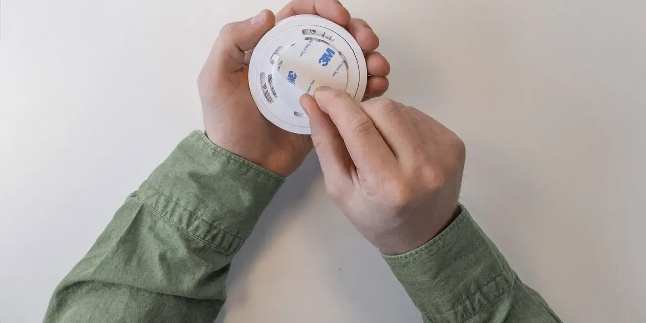Händer drar av skyddstejp från en Verisure brandvarnare, redo att monteras.