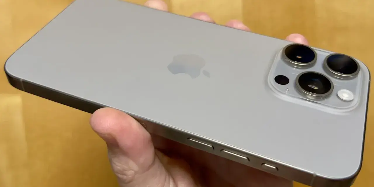En hand håller en silverfärgad iPhone 15 Pro Max. Kameramodulen med tre linser syns tydligt.