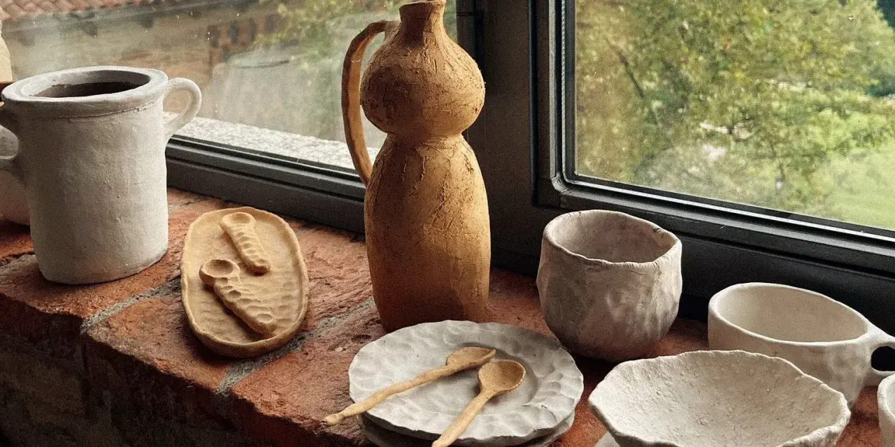 Ceramica fatta in casa: vasi, ciotole e cucchiaini grezzi, posati su un davanzale di mattoni, con vista su un giardino verde.