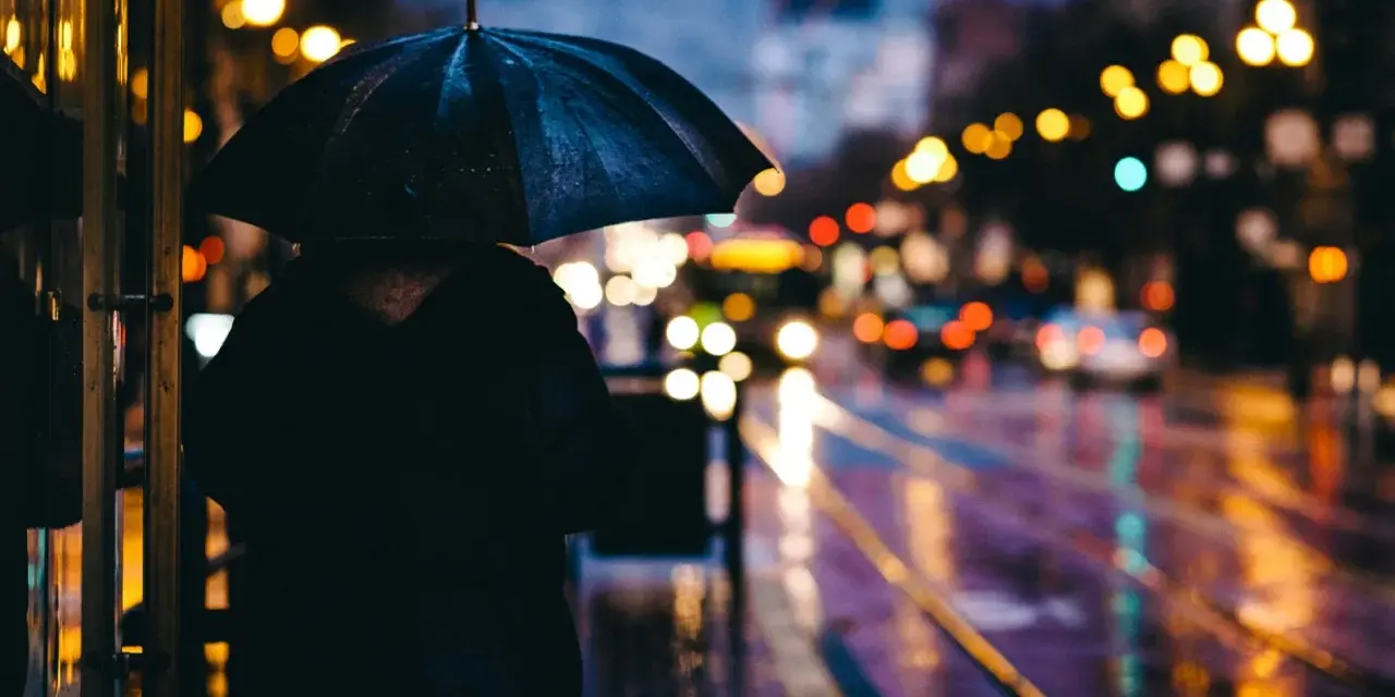 Person sous un parapluie noir, attendant sur un trottoir mouillé. Les lumières de la ville se reflètent sur la chaussée, créant de belles photos de pluie.