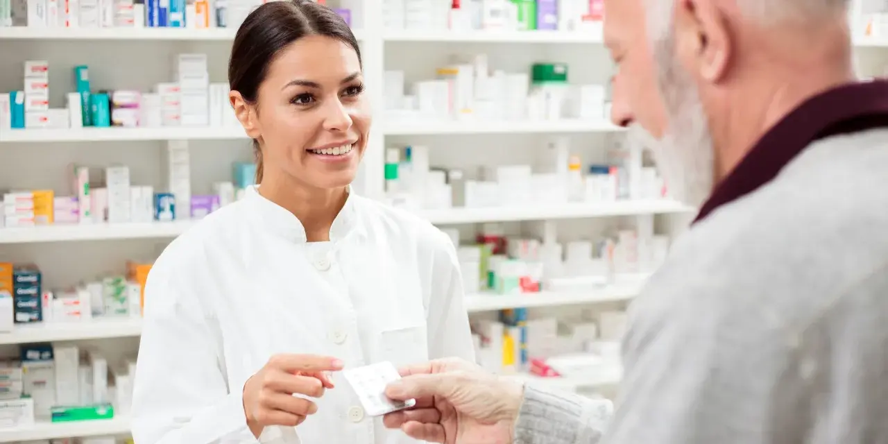 Uśmiechnięta farmaceutka podaje starszemu panu leki. W tle apteczne półki pełne leków, w tym poltixa.