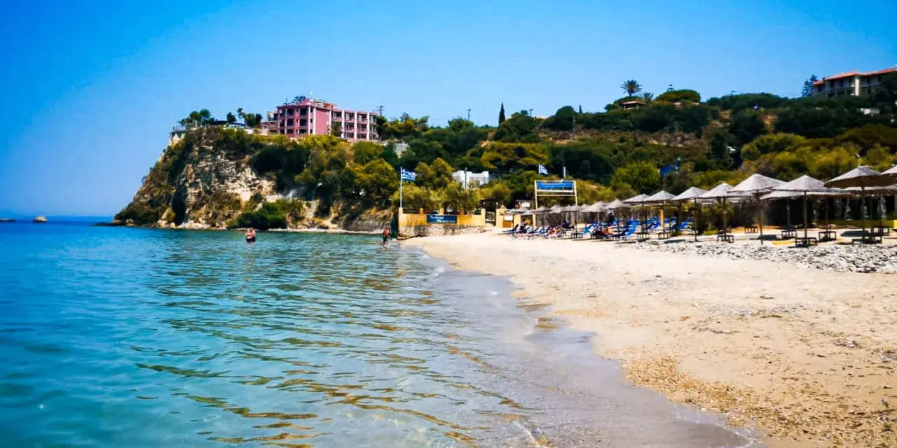 Piękne, turkusowe morze na Zakynthos. Plaża z leżakami i parasolami, a na klifie hotel.