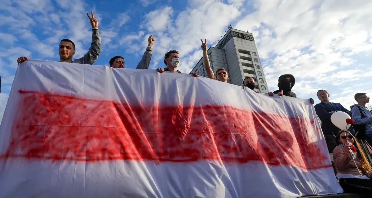 Ludzie trzymają biało-czerwoną flagę, symbol wolności Białorusi. Widać budynek w tle.