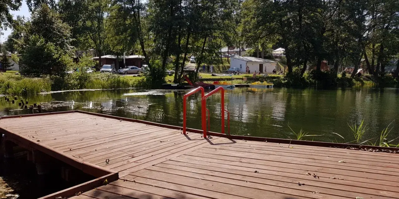 Drewniany pomost z czerwonymi poręczami nad jeziorem. W tle drzewa, budynki i samochody. Idealne miejsce na łagów camping.