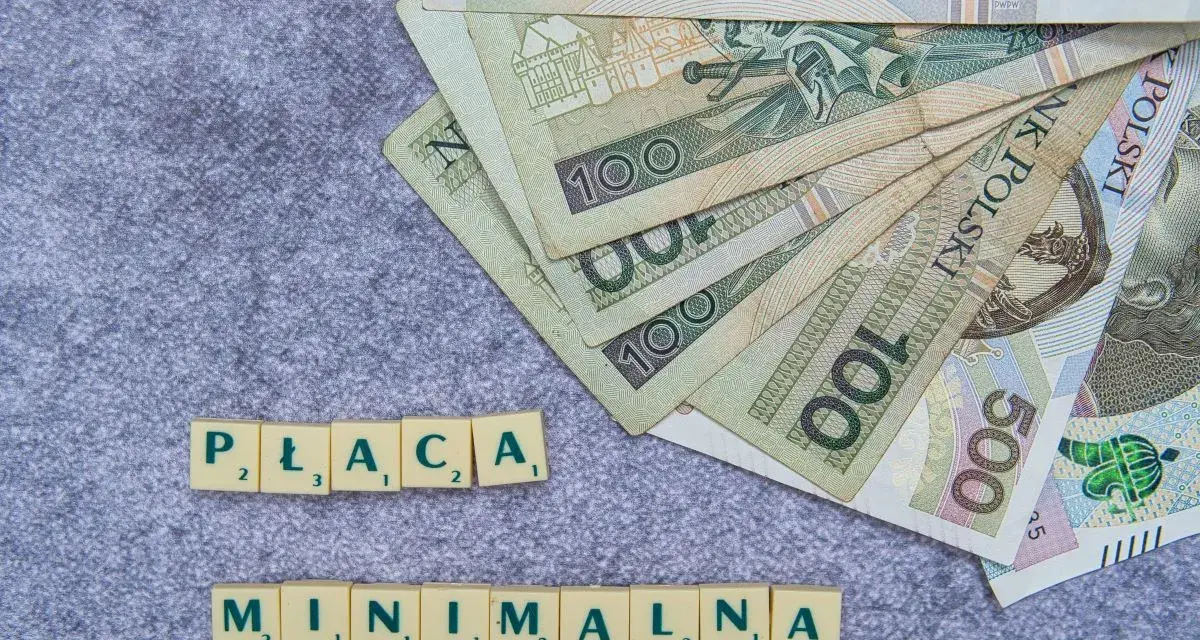 Płaca minimalna w Polsce – polskie banknoty o nominałach 100 i 500 zł ułożone na szarym tle, obok kafelki z literami tworzące napis "PŁACA MINIMALNA".