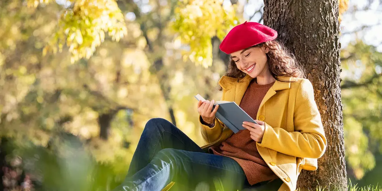 Mujer con boina roja lee un libro bajo un árbol, inspirada por los temas de cuentos del otoño.