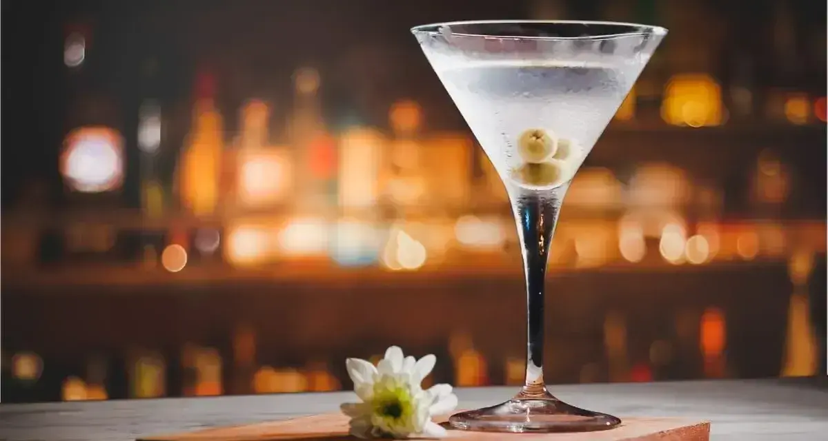 Eleganckie martini z colą, ozdobione oliwkami, na tle rozmytego baru. Obok kieliszka leży biały kwiatek.