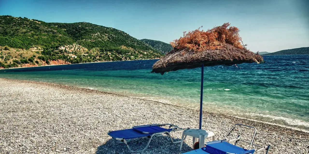 Plaża z leżakami i parasolem, w tle zielone wzgórza i turkusowe morze. To idealne miejsce na wypoczynek na grupie wysp na Morzu Egejskim.