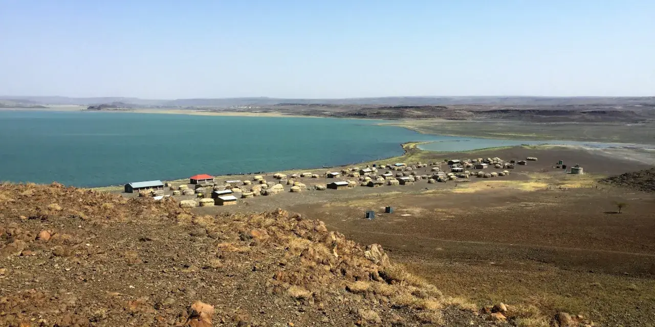 Widok na osadę nad turkusowym jeziorem Turkana. W oddali widać górzysty krajobraz.