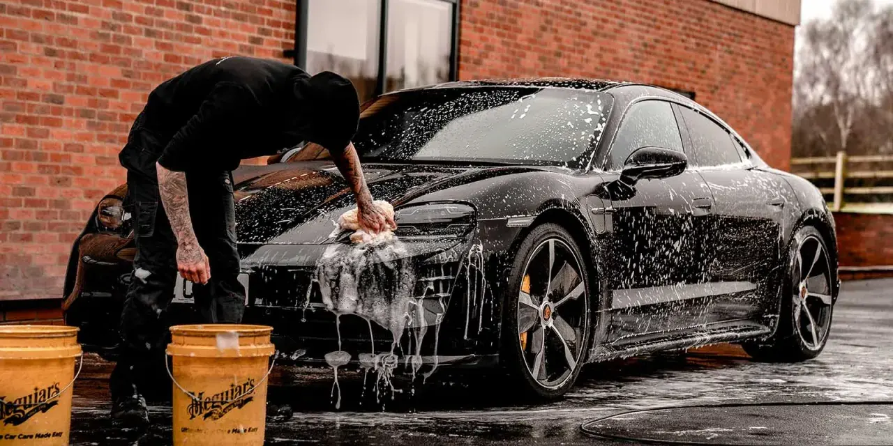 Person tvättar en svart bil med Autoglym-produkter. Två gula hinkar står bredvid.