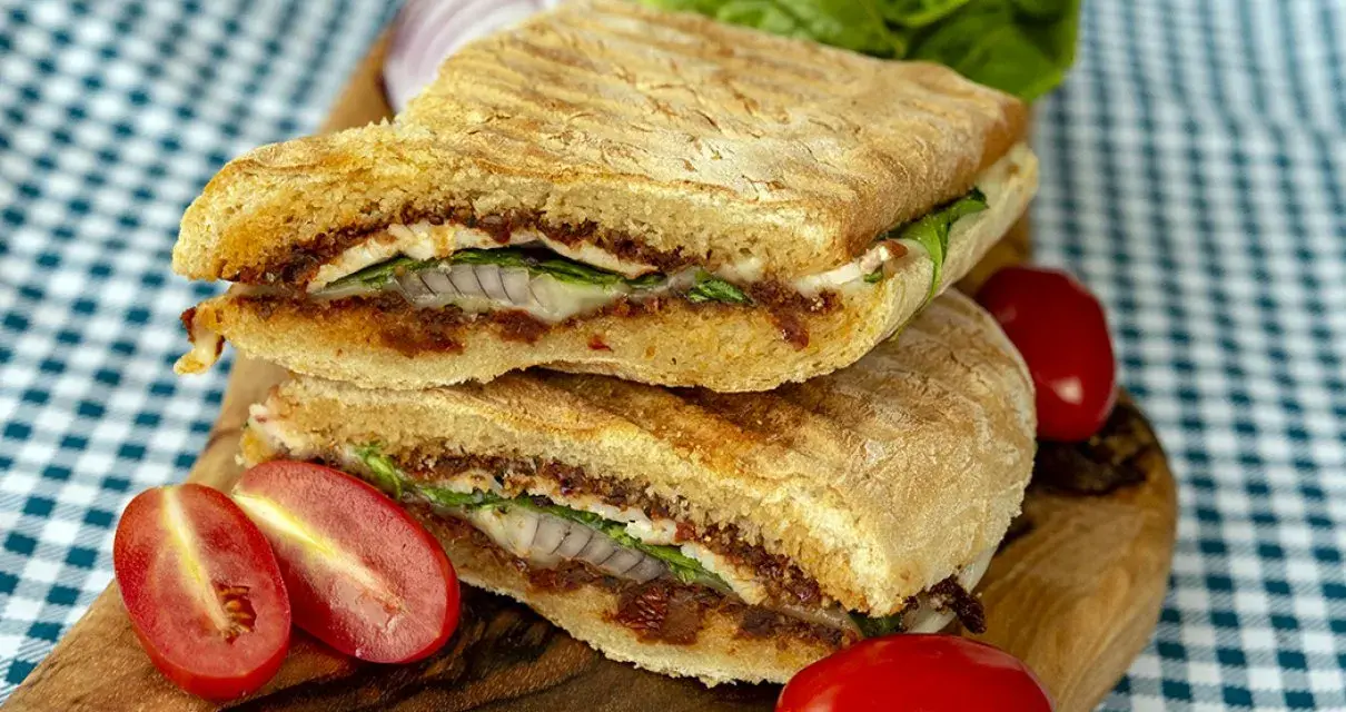 Dwie połówki panini z serem, suszonymi pomidorami, rukolą i cebulą na desce, obok pomidorków i sałaty.