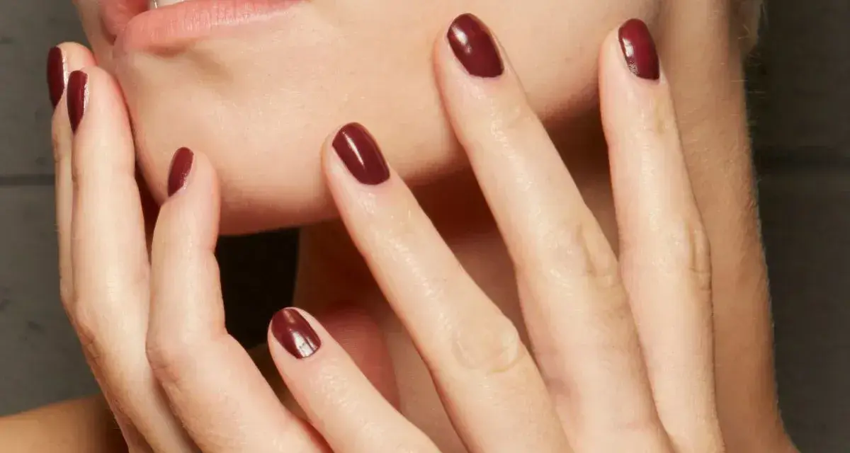 Dłonie z idealnym, ciemnoczerwonym manicurem kombinowanym. Delikatne muśnięcie policzka podkreśla elegancję.