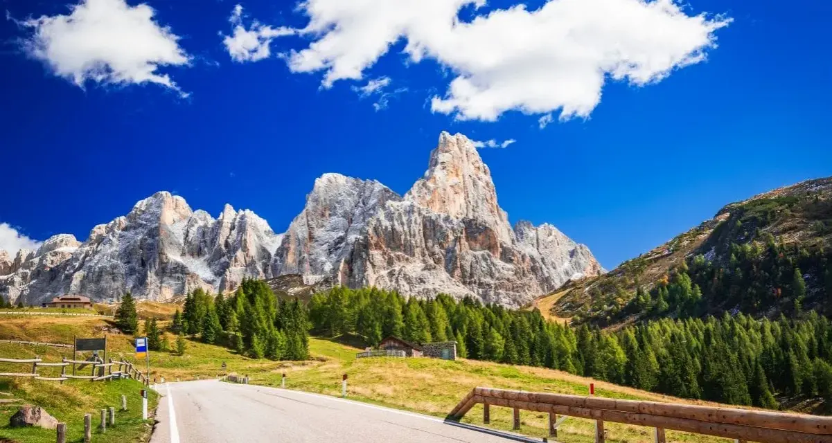 Strada panoramica per esplorare i passi del Trentino in moto, con imponenti vette dolomitiche sullo sfondo e un cielo azzurro con nuvole bianche.