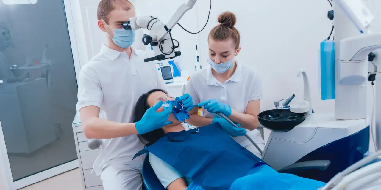 Dentysta pod mikroskopem bada ząb pacjentki, by wykluczyć zatrucie zęba. Asystentka przygotowuje narzędzia.
