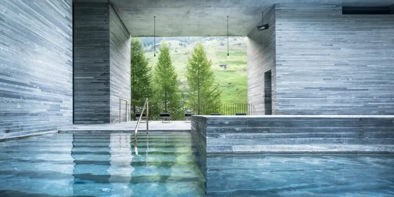 Piscine intérieure des thermes de Vals Zumthor, avec vue sur les montagnes verdoyantes et les sapins. L'eau bleue reflète les murs en pierre grise.