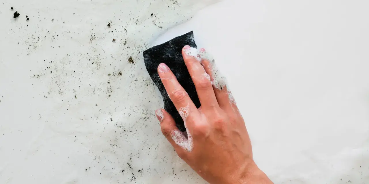 Main d'une personne lavant un mur humide avec une éponge savonneuse. Des saletés et des débris sont visibles sur la surface.