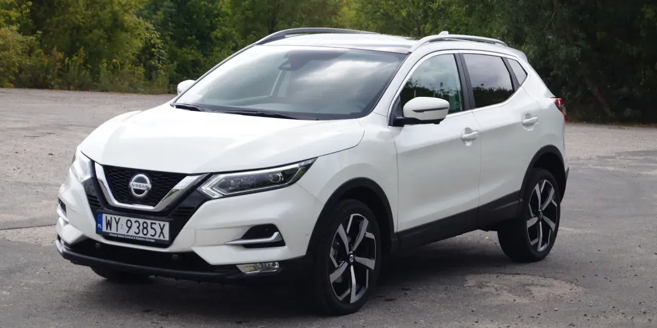 Biały Nissan Qashqai 2014 z polskimi tablicami rejestracyjnymi stoi na drodze, otoczony zielenią drzew.