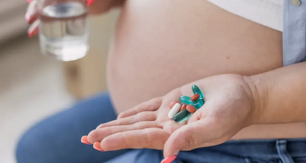Kobieta w ciąży trzyma na dłoni suplementy, w tym żelazo, przygotowując się do ich zażycia z wodą.