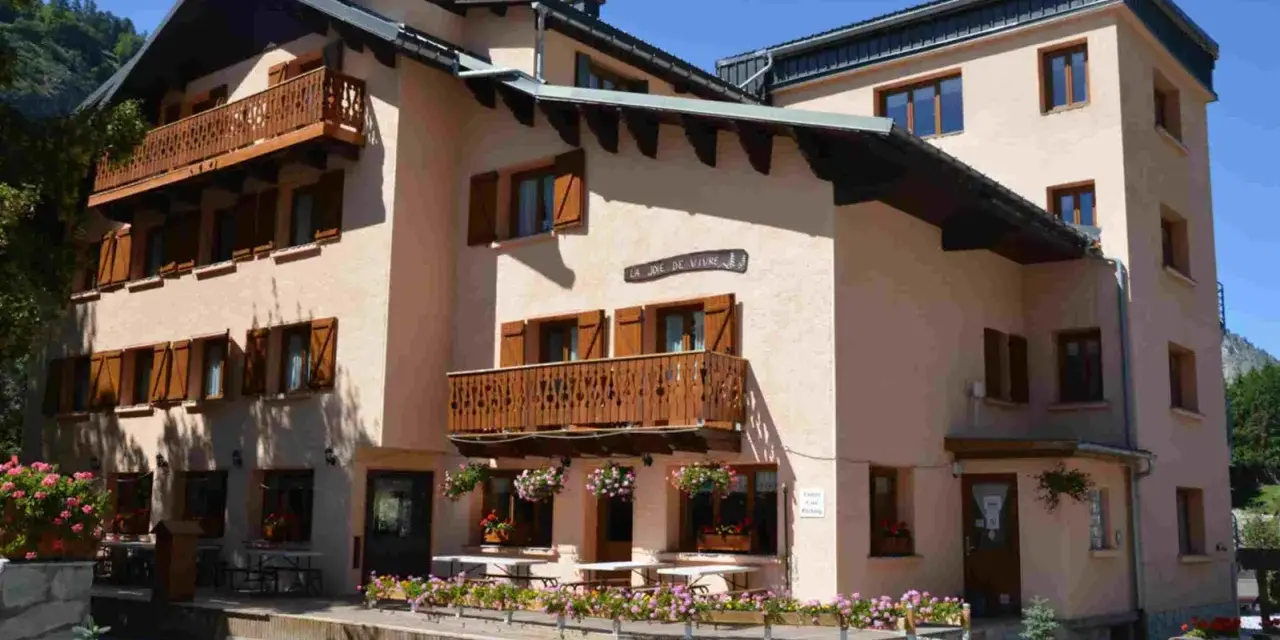 Bâtiment de style alpin avec balcons en bois et volets marrons, fleuri. Une enseigne "La Joie de Vivre" suggère une ambiance de colonie en Haute-Savoie.