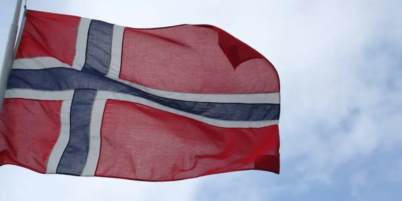 Flaga Norwegii powiewa na wietrze na tle błękitnego nieba z chmurami.
