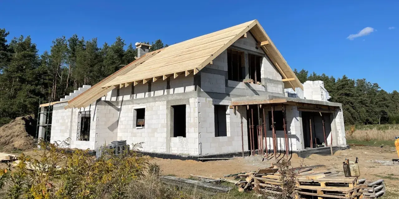 Budowa domu w toku, z widocznym szkieletem dachu i ścianami z bloczków. Warto sprawdzić, ile kosztują formalności przy budowie domu, zanim zaczniemy.