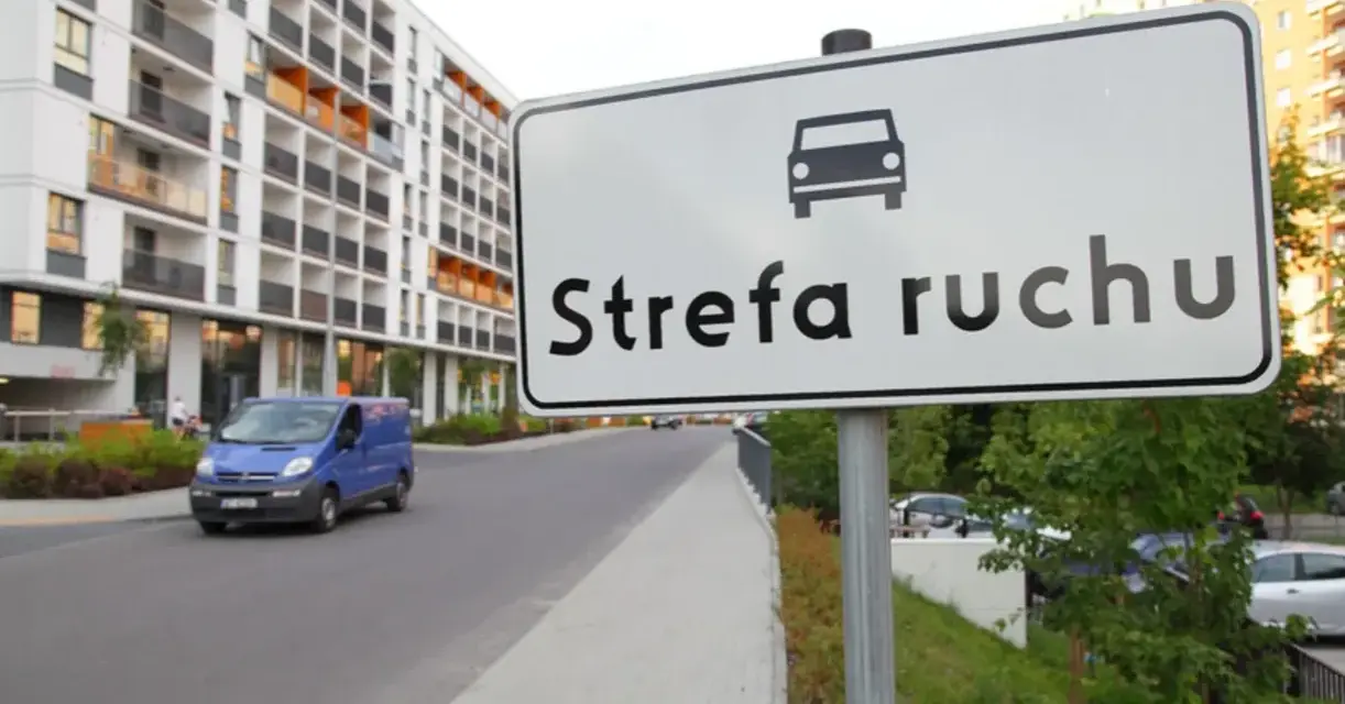 Znak "Strefa ruchu" przed nowoczesnym budynkiem mieszkalnym. Niebieski bus jedzie po drodze.