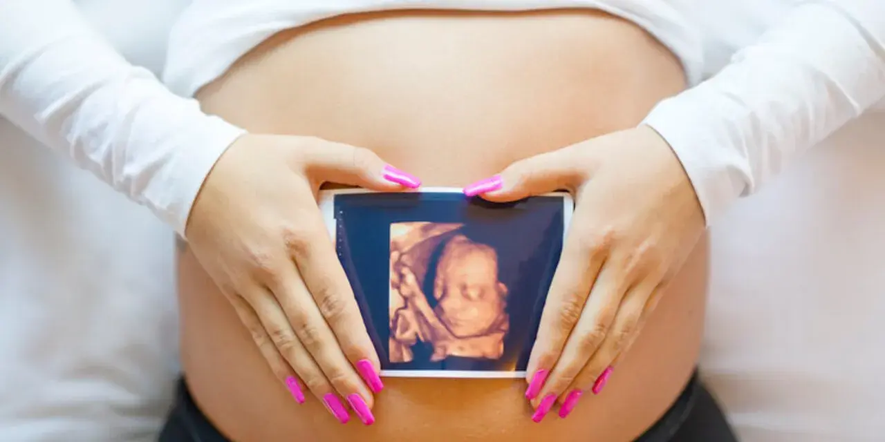 Ręce z różowymi paznokciami obejmują brzuch, na którym widnieje zdjęcie z 8 tygodnia ciąży usg 3d.