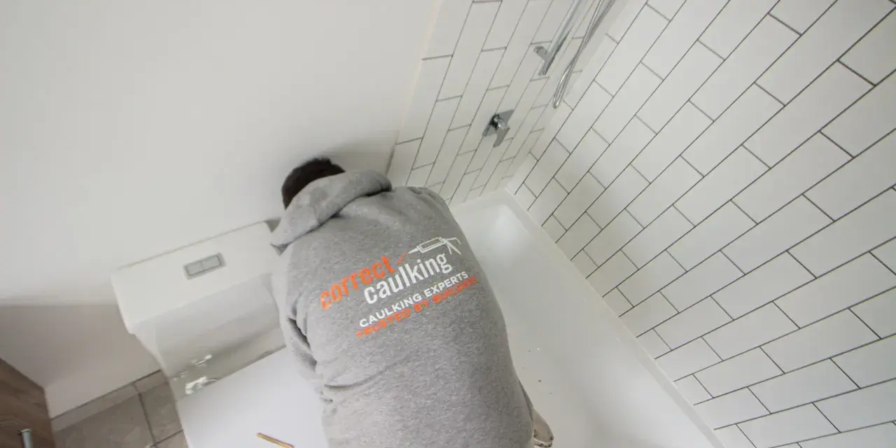 A person in a "Correct Caulking" hoodie works on a bathtub, demonstrating how to caulk a bathtub with white subway tiles in the background.