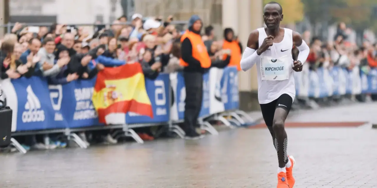 Eliud Kipchoge corre in una maratona, mostrando quanto è lunga una maratona. Pubblico festante e bandiera spagnola sullo sfondo.
