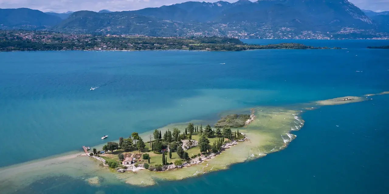 L'isola dei conigli sul Lago di Garda, un gioiello verde con case e alberi, circondata da acque turchesi e montagne sullo sfondo.