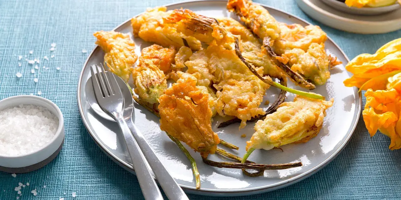 Piatto di fiori di zucca in pastella dorati e croccanti, serviti con sale grosso e posate.