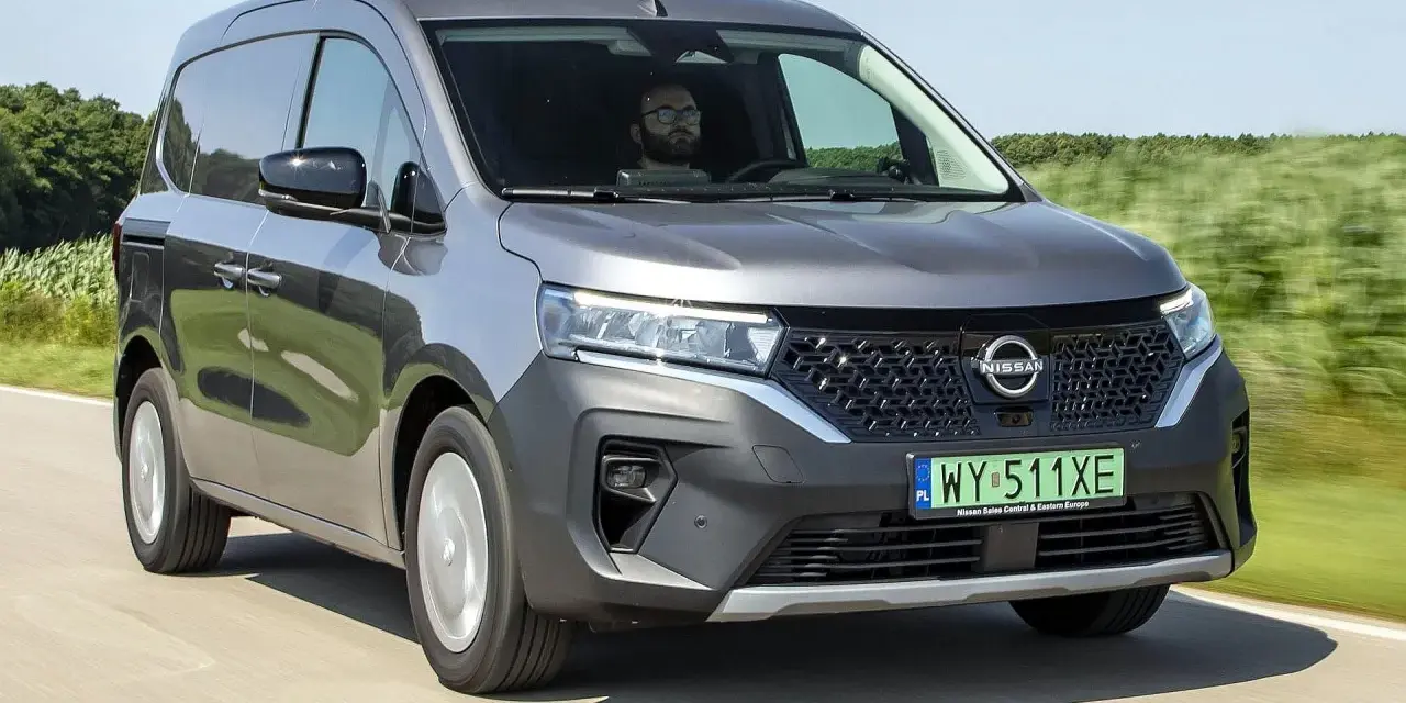 Szary Nissan Townstar jedzie po drodze, otoczony zielonymi polami. Kierowca w okularach prowadzi pojazd.