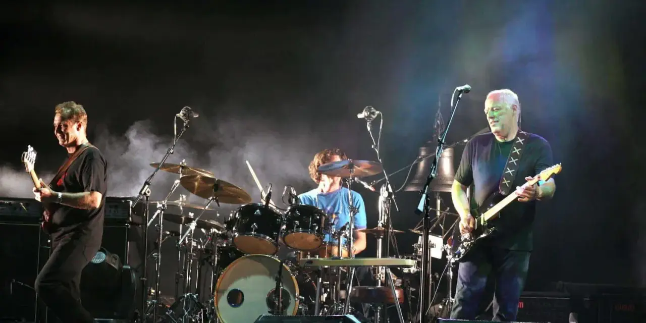 Les membres de Pink Floyd sur scène, David Gilmour à la guitare, Nick Mason à la batterie, et un autre musicien.