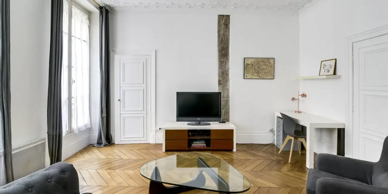 Salon moderne avec parquet en chevrons, canapés gris, bureau et TV. Les murs et le plafond sont peints à l'huile, offrant une finition impeccable.