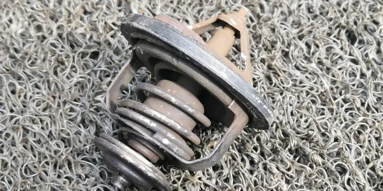 Zużyty termostat w samochodzie, z widocznym mechanizmem sprężynowym i uszczelką, leży na szarej, plecionej powierzchni.
