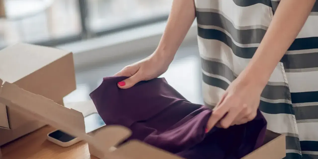 Kobieta pakuje ciemnofioletową sukienkę do kartonu, przygotowując ją do **vinted zwroty**.