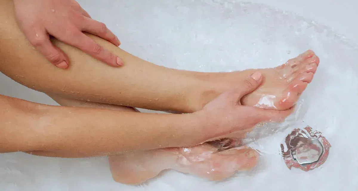 A person's legs and feet are submerged in water, with hands gently massaging the leg. This scene suggests a relaxing foot soak, perhaps to see if epsom salt helps plantar fasciitis.