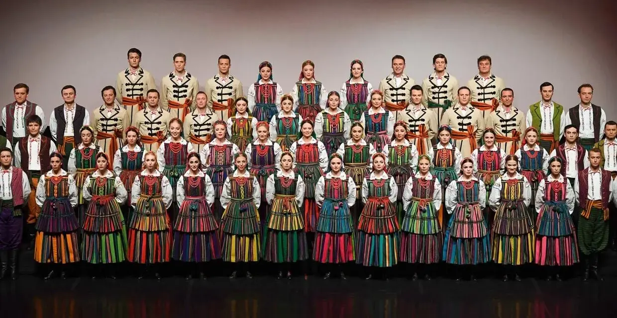 Zespół w barwnych strojach ludowych, prezentujący mazowieckie piosenki ludowe.