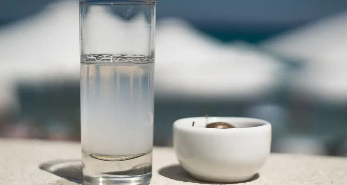 Vaso de pastis bebida, con una aceituna en un cuenco blanco. El fondo está desenfocado, sugiriendo un día soleado.