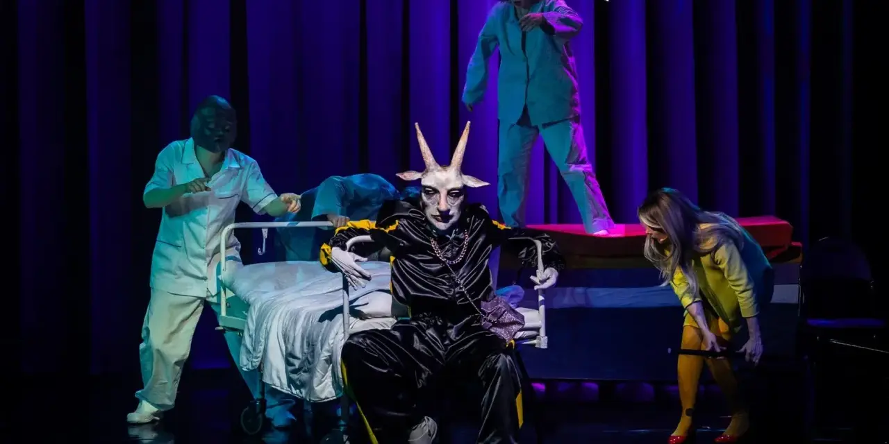 Na scenie, w surrealistycznej atmosferze, panna obsada w stroju z głową kozła siedzi na wózku. Wokół niej, w jasnych fartuchach, poruszają się postacie przypominające personel medyczny.