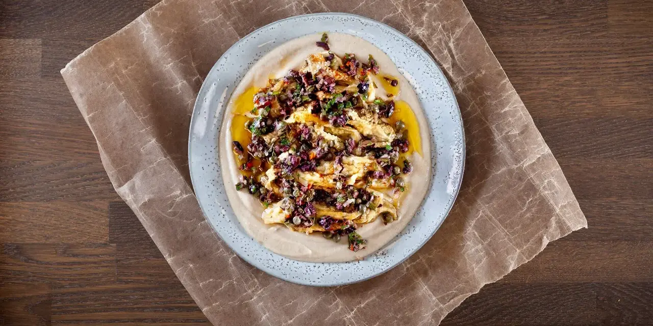 En krämig aubergine i ugn serveras på en bädd av hummus, toppad med en smakrik blandning av oliver, kapris och örter.